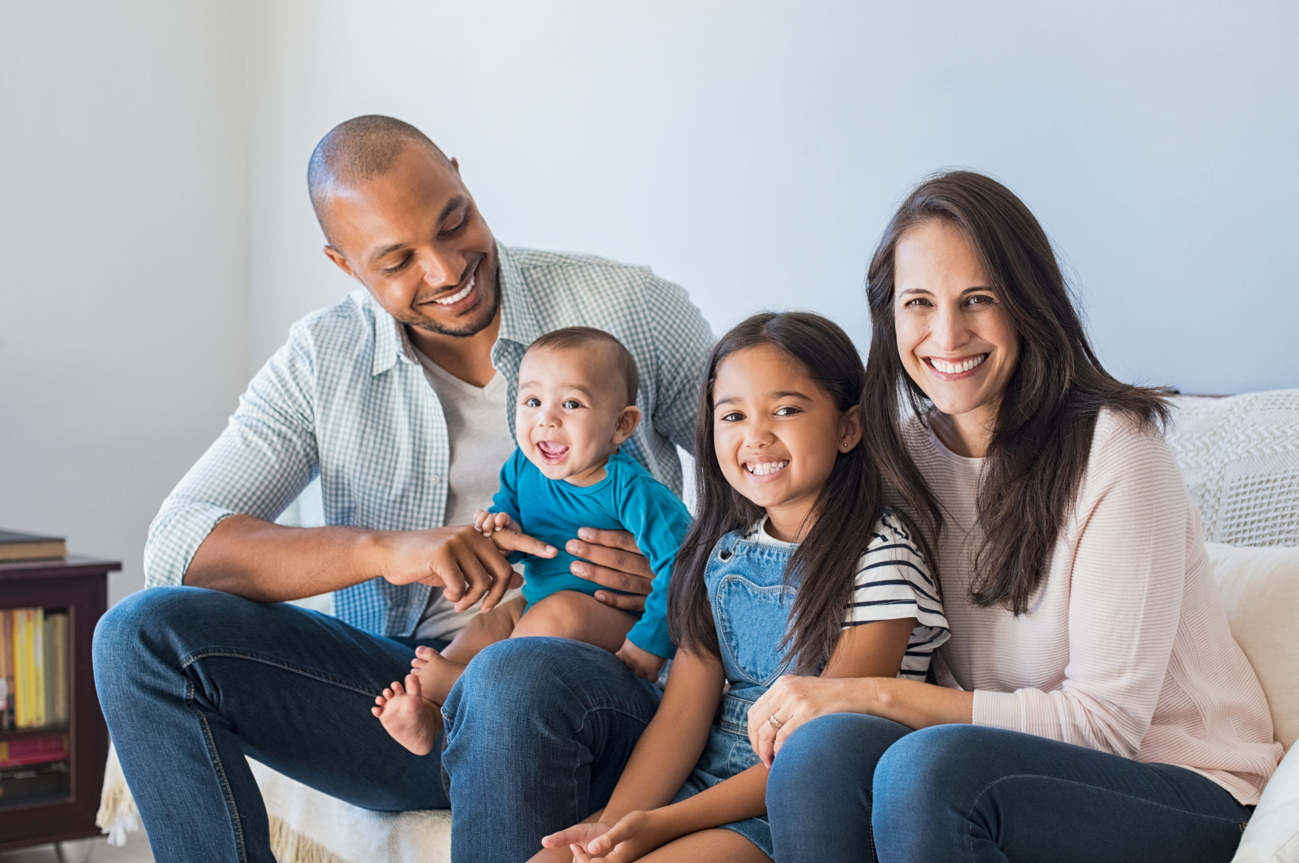 Portrait of happy multiethnic family sitting on sofa at home. Smiling couple with kids sitting on couch and looking at camera. Black father and latin woman with daughter sitting on couch and having fun.