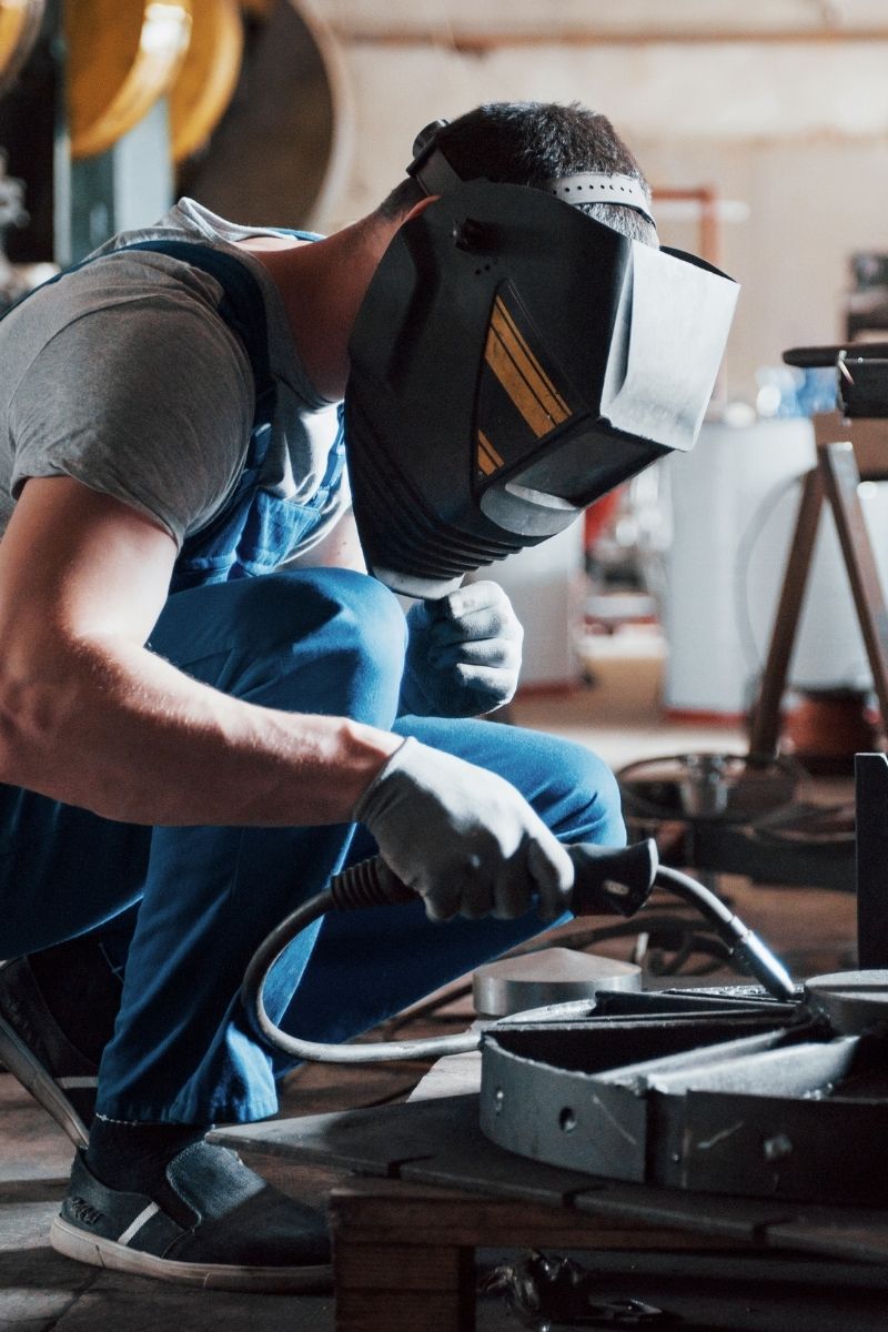 man wearing overalls crouched down by a metal item wearing a welders mask