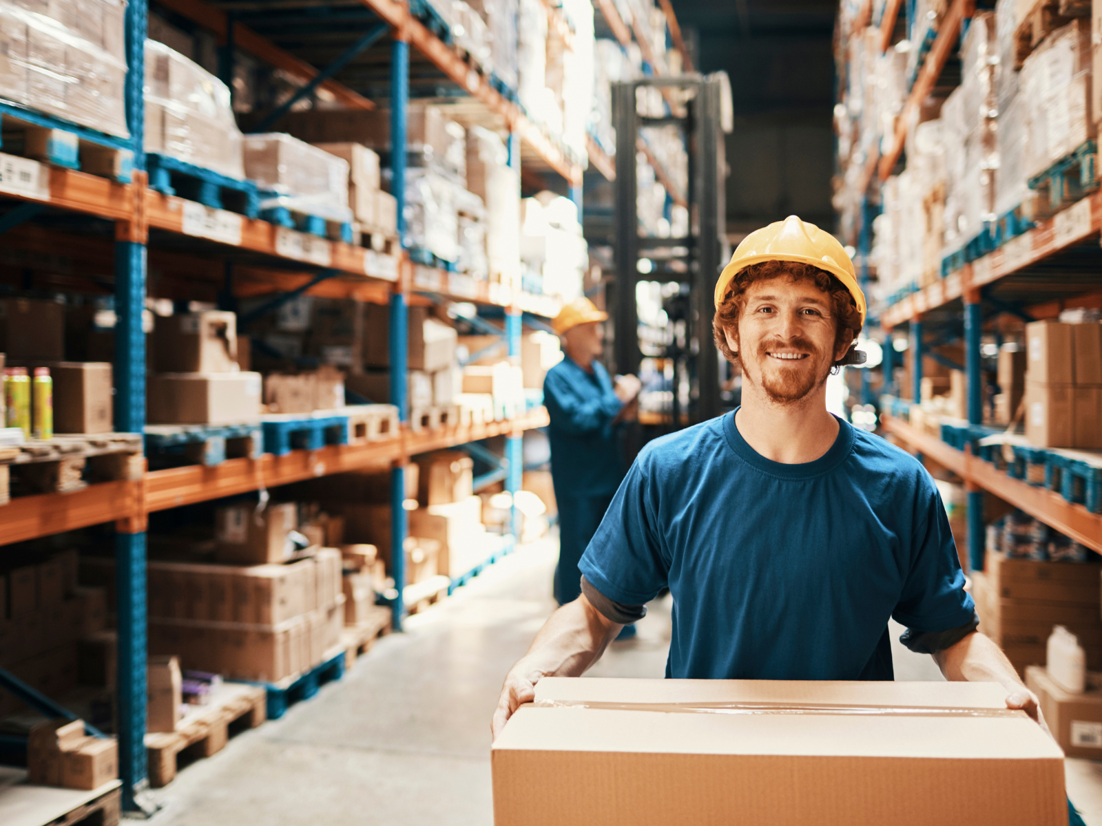 smiling man in a warehouse isle holding a cardboard box surrounded by other boxes on shelves